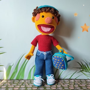 Puede incluir: Una mu&ntilde;eca de crochet con piel amarilla, cabello casta&ntilde;o y una camisa roja. La mu&ntilde;eca lleva pantalones vaqueros azules, una mochila azul y amarilla y zapatillas azules y blancas. La mu&ntilde;eca est&aacute; de pie en un camino de piedra.