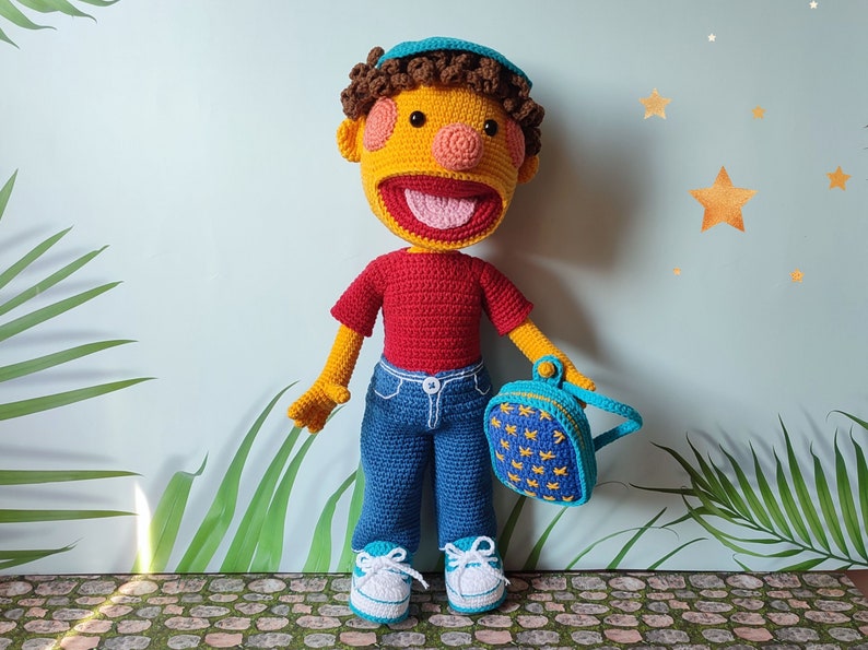 Puede incluir: Una mu&ntilde;eca de crochet con piel amarilla, cabello casta&ntilde;o y una camisa roja. La mu&ntilde;eca lleva pantalones vaqueros azules, una mochila azul y amarilla y zapatillas azules y blancas. La mu&ntilde;eca est&aacute; de pie en un camino de piedra.