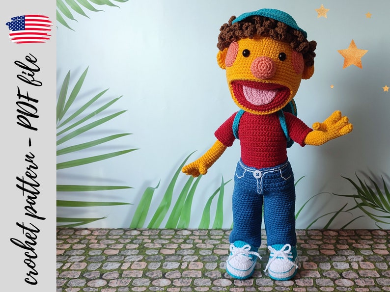 Puede incluir: Juguete de peluche tejido a crochet de un ni&ntilde;o con cabello casta&ntilde;o, que lleva un sombrero azul, una camisa roja, pantalones vaqueros azules y zapatillas blancas. El juguete est&aacute; de pie en un camino de piedra con plantas verdes en el fondo.