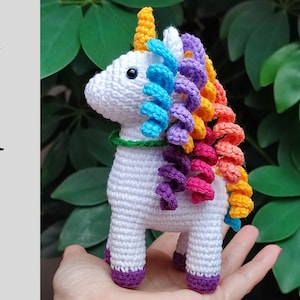 May include: A white crocheted unicorn with a rainbow mane. The unicorn has a green collar and purple hooves.