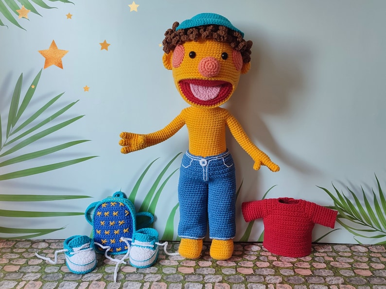 Puede incluir: Una mu&ntilde;eca de crochet con piel amarilla, cabello casta&ntilde;o y un sombrero azul. La mu&ntilde;eca lleva pantalones vaqueros azules y una camiseta roja. La mu&ntilde;eca lleva una mochila azul y un par de zapatos azules.