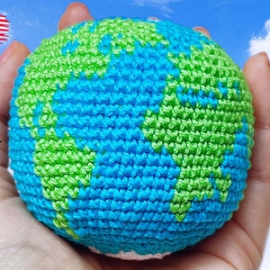 May include: A crocheted globe with blue and green yarn, representing the Earth, held in a hand against a blue sky with white clouds.
