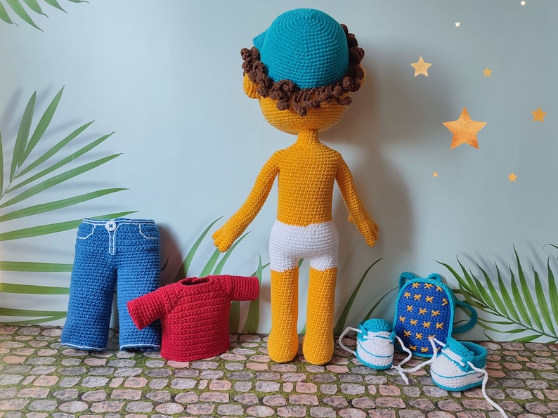 Puede incluir: Una mu&ntilde;eca de crochet con cabello casta&ntilde;o que lleva un sombrero azul, pantalones cortos blancos y zapatos azules. La mu&ntilde;eca est&aacute; de pie sobre una superficie de piedra con una camisa roja, pantalones vaqueros azules y una mochila azul con estrellas amarillas.