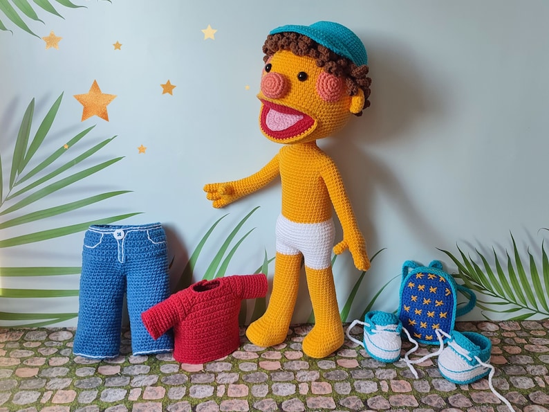Puede incluir: Un t&iacute;tere amarillo de crochet con un sombrero azul, boca roja y pelo marr&oacute;n. El t&iacute;tere lleva pantalones cortos blancos y est&aacute; de pie sobre una superficie de piedra. En la imagen hay unos vaqueros azules, una camiseta roja, una mochila azul con estrellas amarillas y dos pares de zapatillas azules y blancas.