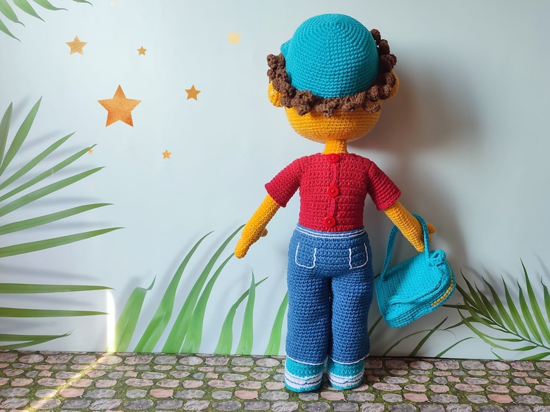 Puede incluir: Una mu&ntilde;eca de crochet con un sombrero azul, una camisa roja, pantalones vaqueros azules y llevando un bolso azul. La mu&ntilde;eca tiene el pelo marr&oacute;n y la cara amarilla.