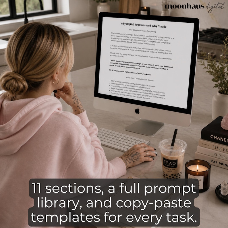 Woman in a pink hoodie sitting at a desktop computer reading a prompt engineering guide about creating digital products with Claude AI. Overlay text reads “11 sections, a full prompt library, and copy-paste templates for every task” while the workspace includes candles, books, and bubble tea beside the computer.