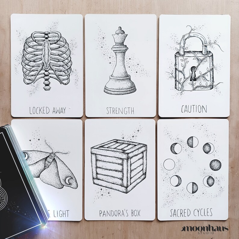 6 cards from the wandering soul oracle deck, showing moon phases, lunar moth, antique lock, rib cage, the queen chess piece, wooden sealed crate. drawn in black and white. indie tarot oracle deck.