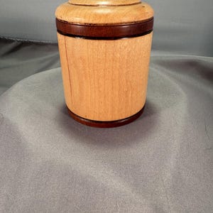 May include: A cylindrical wooden container featuring a light tan body with a dark brown base and a matching dark brown lid. The lid has a smooth, rounded top section and a flat, dark brown rim that sits flush against the light tan main body.