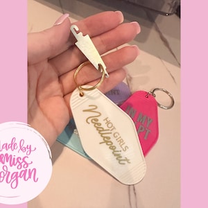 May include: White motel key chain with gold lettering that says "Hot Girls Needlepoint". A pink motel key chain with silver lettering that says "In My Apt" is behind the white key chain.