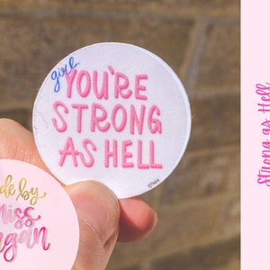 May include: A pink and white round sticker with the text "girl you're strong as hell" in pink lettering. The sticker is held in a hand. The sticker is made by "miss morgan" and is labeled "strong as hell sticker".
