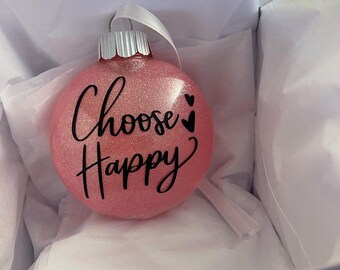 May include: A pink glitter Christmas ornament with the words "Choose Happy" in black script. The ornament has a silver cap and a white ribbon for hanging. It is presented in a white box with tissue paper.