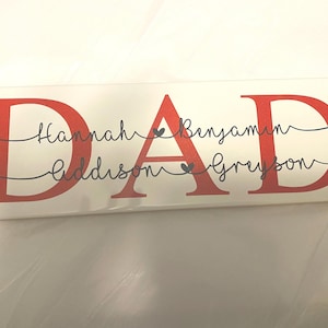 May include: A rectangular white sign with the word "DAD" in large red letters. Below the letters are the names Hannah, Addison, Benjamin, and Greyson in black script. Small heart symbols are between the names.