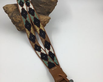 May include: A beaded strap with a geometric pattern in black, white, brown, and blue. The strap has a leather loop and a brown leather tassel.