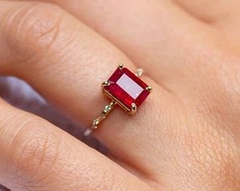 Op de afbeelding: Een gouden ring met een rechthoekige, dieprode edelsteen. De band van de ring is versierd met kleine, delicate details. De ring wordt op een hand getoond, waarbij het ontwerp en de kleur van het sieraad worden benadrukt.