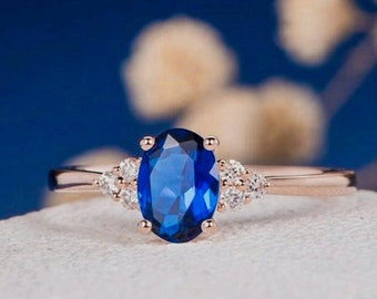 May include: A rose gold ring featuring a large, oval-cut, deep blue gemstone as the centerpiece. Flanking the central stone are smaller, clear accent stones. The ring is set against a soft, white surface with a blurred blue background.