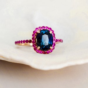 May include: A gold ring featuring a large, dark blue gemstone surrounded by a halo of smaller, pink gemstones. The ring's band is also adorned with pink gemstones. The ring is set against a white background.