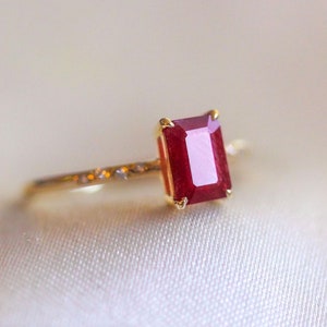 Può includere: Un anello d'oro con una grande gemma rettangolare rosso scuro. La fascia dell'anello è delicata, con piccoli abbellimenti. La gemma è incastonata in una lunetta d'oro, creando un look classico ed elegante.