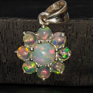 May include: A silver pendant featuring a flower design with a central opal surrounded by eight smaller opals. The opals display iridescent colors of green, pink, and yellow. The pendant has a loop for a chain.