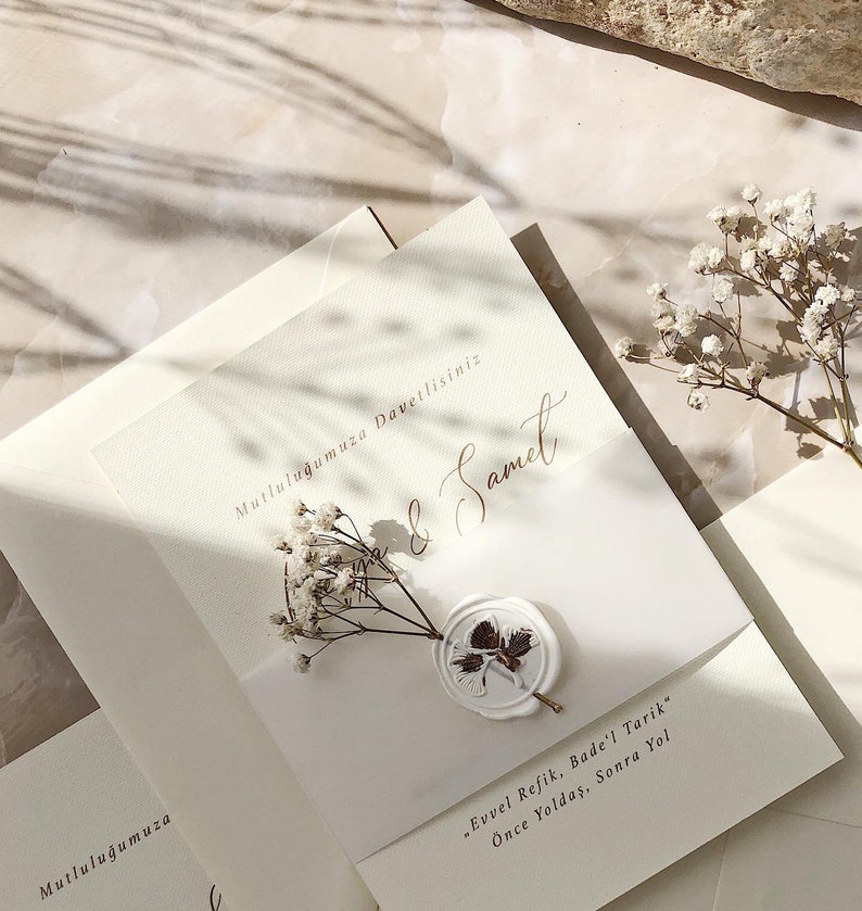 May include: A white wedding invitation with a wax seal and a sprig of white baby's breath. The invitation reads "Mutluluğumuza Davetlisiniz" and "& Samet". The text below the seal reads "Evvel Refik, Bade'l Tarik" and "&Ouml;nce Yoldaş, Sonra Yol".