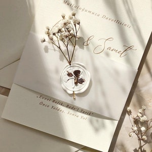 May include: A white wedding invitation with a wax seal featuring a brown floral design. The invitation is decorated with white baby's breath flowers and includes the text "Mutluluğumuza Davetlisiniz" and "& Samet". The text "Evvel Refik, Bade'l Tarik" and "&Ouml;nce Yoldaş, Sonra Yol" is also included.