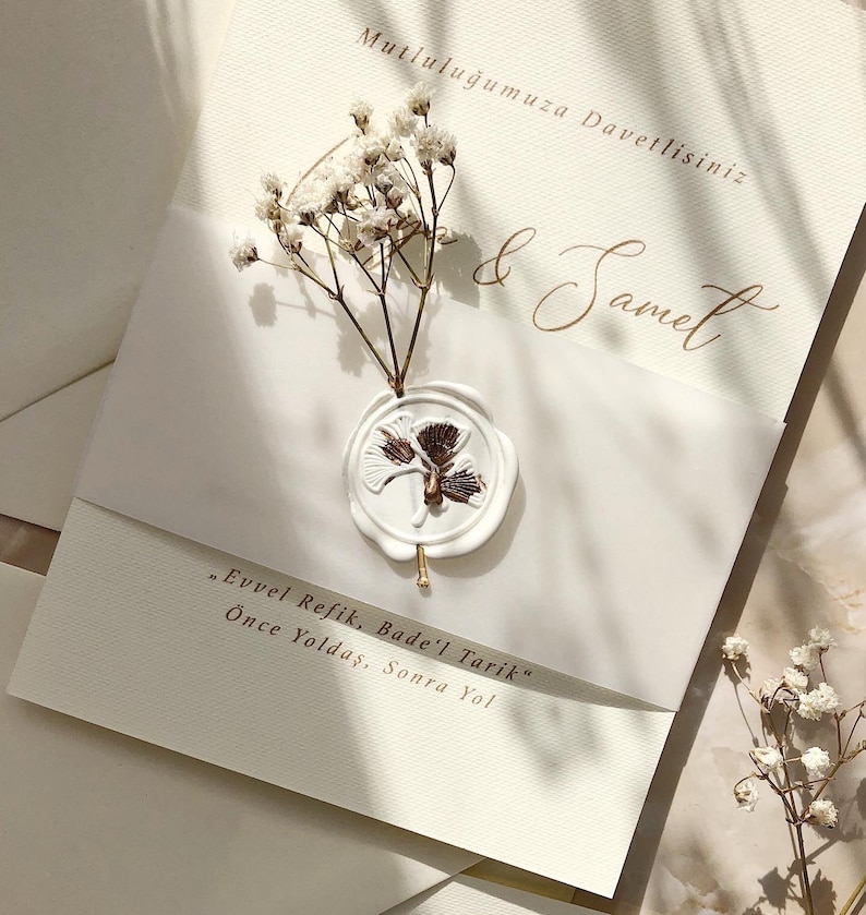May include: A white wedding invitation with a wax seal featuring a brown floral design. The invitation is decorated with white baby's breath flowers and includes the text "Mutluluğumuza Davetlisiniz" and "& Samet". The text "Evvel Refik, Bade'l Tarik" and "&Ouml;nce Yoldaş, Sonra Yol" is also included.
