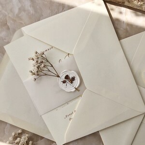 May include: A white envelope with a wax seal and a small bouquet of white flowers. The envelope is folded in half and has a brown inscription that reads "Evvel P" and "Or".