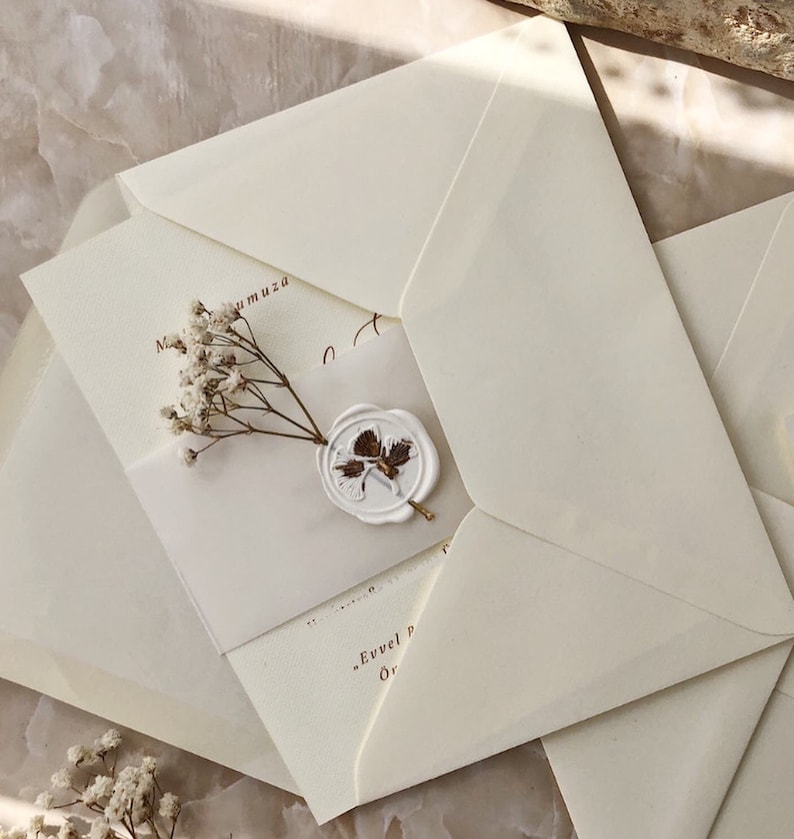 May include: A white envelope with a wax seal and a small bouquet of white flowers. The envelope is folded in half and has a brown inscription that reads "Evvel P" and "Or".