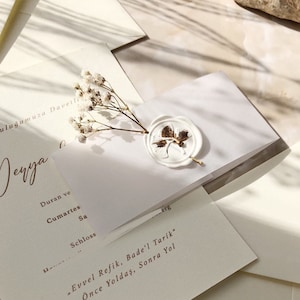 May include: A white envelope with a wax seal featuring a gold leaf design. The envelope is decorated with small white flowers and has the text "Evvel Refik, Bade'l Tarik" and "&Ouml;nce Yoldaş, Sonra Yol" written in gold.