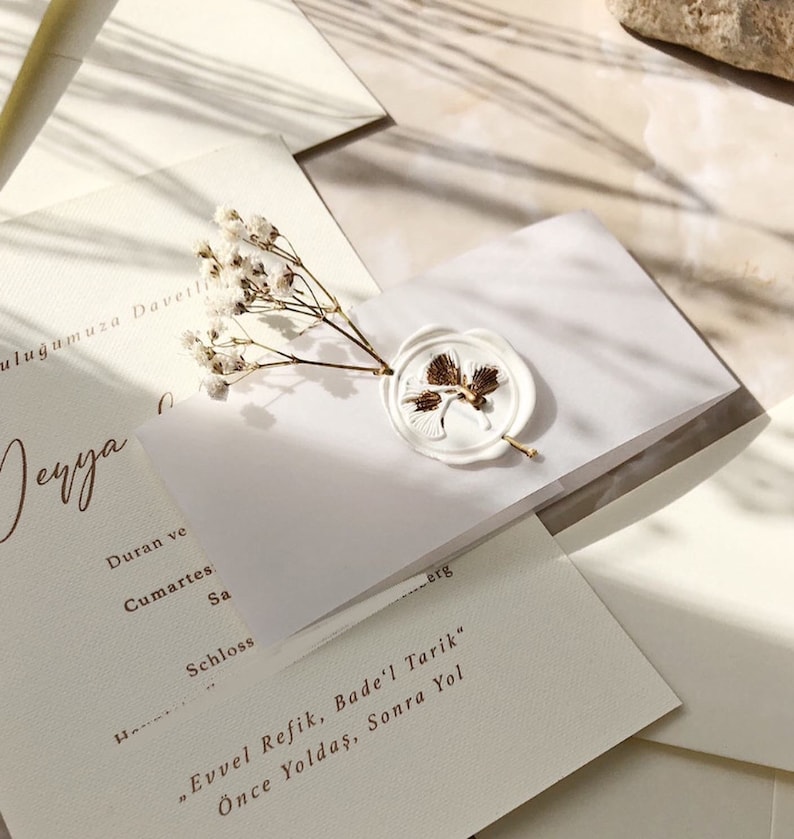 May include: A white envelope with a wax seal featuring a gold leaf design. The envelope is decorated with small white flowers and has the text "Evvel Refik, Bade'l Tarik" and "&Ouml;nce Yoldaş, Sonra Yol" written in gold.