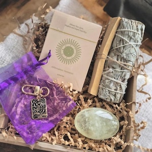 May include: A gift box with a sage bundle, a pale green crystal, a card deck with a sun design, and a purple pouch with a keychain. The card deck reads "Affirmation Moments". The items are in brown paper filler.