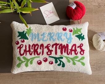 Puede incluir: Una almohada blanca con las palabras "Merry Christmas" bordadas en hilo rojo, azul y verde. La almohada está rodeada de hojas verdes y bayas rojas.