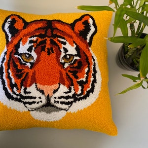 Puede incluir: Una almohada amarilla con un rostro de tigre bordado. El tigre tiene rayas naranjas y negras y pelaje blanco alrededor de la nariz.