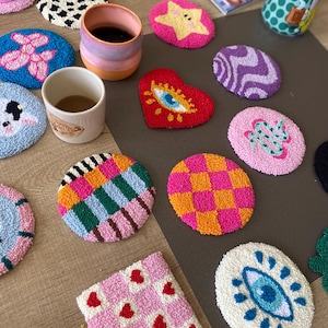 Puede incluir: Una colección de posavasos de punch needle coloridos con varios diseños, incluyendo una estrella amarilla, un corazón rojo con un ojo, un tablero de ajedrez rosa y naranja, una cuadrícula de corazones blancos y rosas y un perro globo azul.