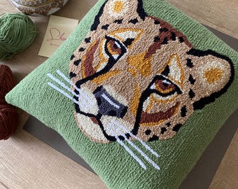 May include: A square, green decorative pillow featuring a detailed embroidered cheetah face. The cheetah's face is rendered in shades of brown, tan, and black, with white whiskers. The pillow is placed on a wooden surface.