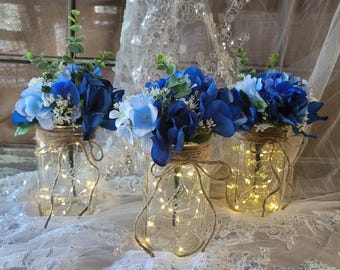 May include: Three mason jars with blue and white flowers and string lights inside. The jars are sitting on a white lace tablecloth.