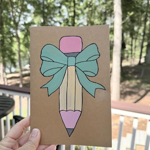 May include: A hand-painted greeting card featuring a pencil design with a pink eraser and tip, a light yellow body, and a teal bow. The card is on a brown background.