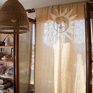 May include: Beige linen curtain featuring a sun design. The sun is embroidered in a lighter beige shade. A woven pendant light hangs from the ceiling. A wooden cabinet and shelves are visible in the background.