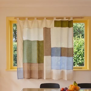May include: A window curtain featuring a colour-blocked patchwork design in cream, olive green, brown, and light blue. The curtain is hung from a wooden rod using fabric tabs and displays a rectangular grid pattern of varied fabric panels.