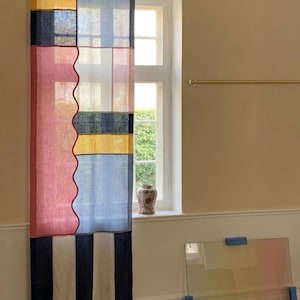 Puede incluir: Una cortina estampada y colorida con un diseño de patchwork. La cortina presenta bloques de tela amarilla, azul, rosa y azul marino, con un borde ondulado. La sección inferior tiene rayas verticales blancas y azul marino. La cortina cuelga frente a una ventana.