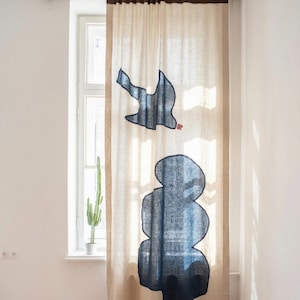 Puede incluir: Cortina de lino beige con un diseño de pájaro azul oscuro y formas abstractas. La cortina cuelga frente a una ventana, con un cactus visible en el fondo. La parte superior de la cortina es de color marrón oscuro.