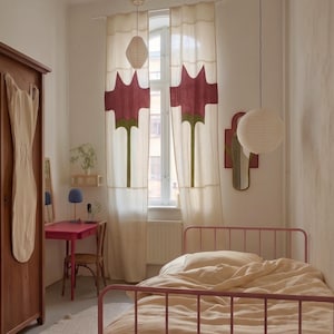 Puede incluir: Interior de dormitorio con una cama de metal rosa y ropa de cama crema. La habitación incluye un armario de madera, un pequeño escritorio rosa y cortinas con un diseño floral. Faroles de papel cuelgan del techo y un espejo está montado en la pared.