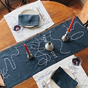 May include: A dark blue linen table runner with white embroidered lines and the text "несовершенство" and "редкость" in Russian. Two place settings are on a wooden table with white linen placemats, dark blue linen napkins, and silver cutlery.