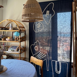 Könnte beinhalten: Esszimmer-Szene mit einer gewebten Pendelleuchte aus Rattan, einer runden blau karierten Tischdecke und einem Holzregal mit dekorativen Gegenständen. Im Hintergrund hängt ein marineblauer Vorhang mit weißen abstrakten Designs.