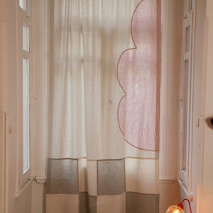 Può includere: Un soggiorno con una grande finestra con una tenda trasparente con un designo a nuvola rosa. La parte inferiore della tenda ha un motivo a scacchi beige e bianco. Una poltrona e un poggiapiedi in pelle marrone sono in primo piano. Una lampada rossa con un globo di vetro trasparente è su un piccolo tavolo di legno.