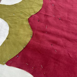 K&ouml;nnte beinhalten: Ein olivgr&uuml;nes Stoffst&uuml;ck mit geschwungener Kante ist auf ein gr&ouml;&szlig;eres St&uuml;ck dunkelroten Stoff gesteckt. Zahlreiche Stecknadeln mit bunten K&ouml;pfen sind &uuml;ber beide Stoffteile verteilt, um die beiden St&uuml;cke sicher miteinander zu verbinden.