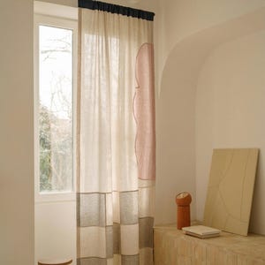 May include: Curtain with a beige and pink pattern, hanging in front of a window. The curtain has a dark blue header and a geometric design. A small wooden stool is in front of the window.