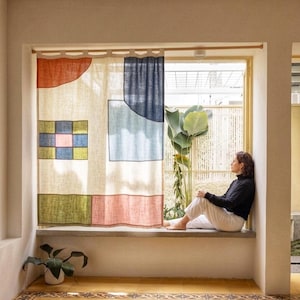 May include: A rectangular window curtain made of off-white fabric featuring a geometric pattern of a red semi-circle, a large blue square, a green and blue grid block, and a pink rectangle, suspended from a wooden rod with fabric loops.