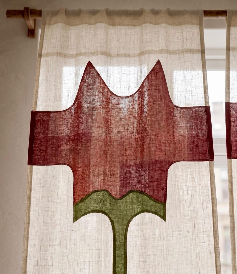 K&ouml;nnte beinhalten: Ein halbtransparenter, cremewei&szlig;er Vorhang an einer Holzstange mit einem gro&szlig;en, mittig applizierten Design einer stilisierten roten Blume mit gr&uuml;nem Stiel und Bl&auml;ttern, gefertigt aus strukturiertem Stoff mit sichtbarem Webmuster.