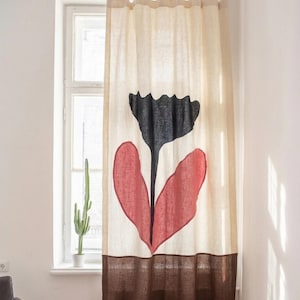 Op de afbeelding: Een beige linnen gordijn met een groot bloemmotief. De bloem heeft een zwarte, geschulpte bovenkant en twee rode hartvormige bladeren. Het onderste derde deel van het gordijn is bruin. Een cactus in een witte pot staat op de vensterbank.