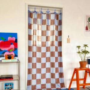 Peut inclure: Un rideau de porte rectangulaire présentant un motif à carreaux bleu clair et marron. Le rideau est suspendu à une tringle blanche par six attaches en tissu bleu clair nouées sur le dessus, créant un habillage de porte simple et fonctionnel.